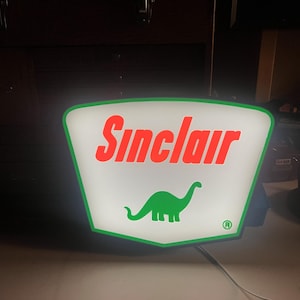 May include: A vintage Sinclair petrol station sign. The sign is a white, shield-shaped design with a green border. The word "Sinclair" is in red, and a green dinosaur is below the text.