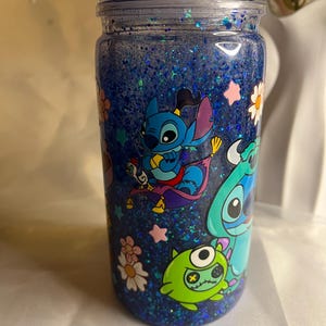 May include: A blue glitter tumbler featuring cartoon characters. The tumbler has a clear lid with small figurines on top. The design includes images of Stitch and other characters, along with flowers and stars. The tumbler is filled with blue glitter.