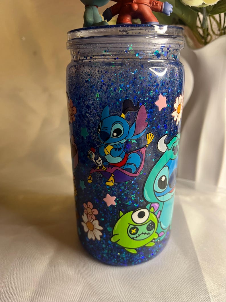 May include: A blue glitter tumbler featuring cartoon characters. The tumbler has a clear lid with small figurines on top. The design includes images of Stitch and other characters, along with flowers and stars. The tumbler is filled with blue glitter.