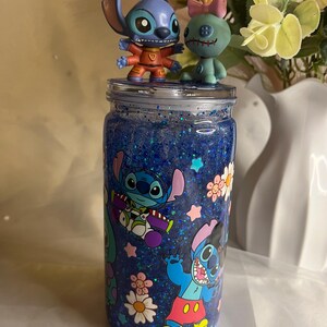 May include: A blue glitter jar with Stitch character decals and two Stitch figurines on top. The jar features a blue glitter interior and cartoon images of Stitch, flowers, and stars. The figurines are a blue Stitch and a green Stitch-like character.