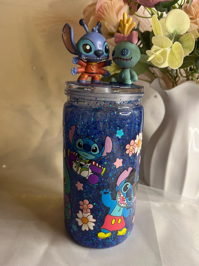 May include: A blue glitter jar with Stitch character decals and two Stitch figurines on top. The jar features a blue glitter interior and cartoon images of Stitch, flowers, and stars. The figurines are a blue Stitch and a green Stitch-like character.
