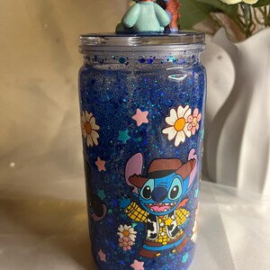 May include: A blue glitter tumbler with a clear lid, featuring a cartoon character in a cowboy outfit. The tumbler is decorated with white daisies and pink stars. A small figurine of a blue alien character sits on top.