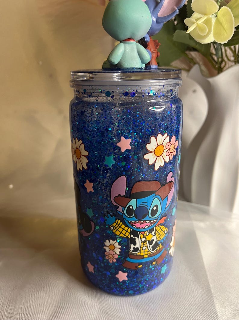 May include: A blue glitter tumbler with a clear lid, featuring a cartoon character in a cowboy outfit. The tumbler is decorated with white daisies and pink stars. A small figurine of a blue alien character sits on top.