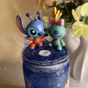 May include: A blue glitter tumbler featuring cartoon characters Stitch and Angel. The tumbler has a blue glitter interior and is topped with two character figurines. The design includes floral and character graphics.