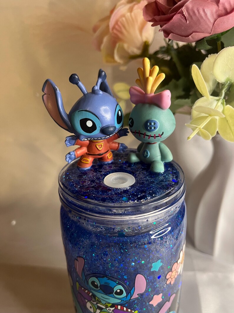 May include: A blue glitter tumbler featuring cartoon characters Stitch and Angel. The tumbler has a blue glitter interior and is topped with two character figurines. The design includes floral and character graphics.