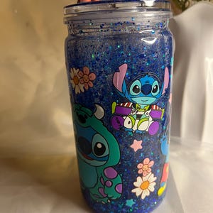 May include: A blue glitter tumbler featuring cartoon characters Stitch, Sulley, and Buzz Lightyear. The tumbler has a clear lid and is decorated with flowers and stars. Two character figurines sit on top.