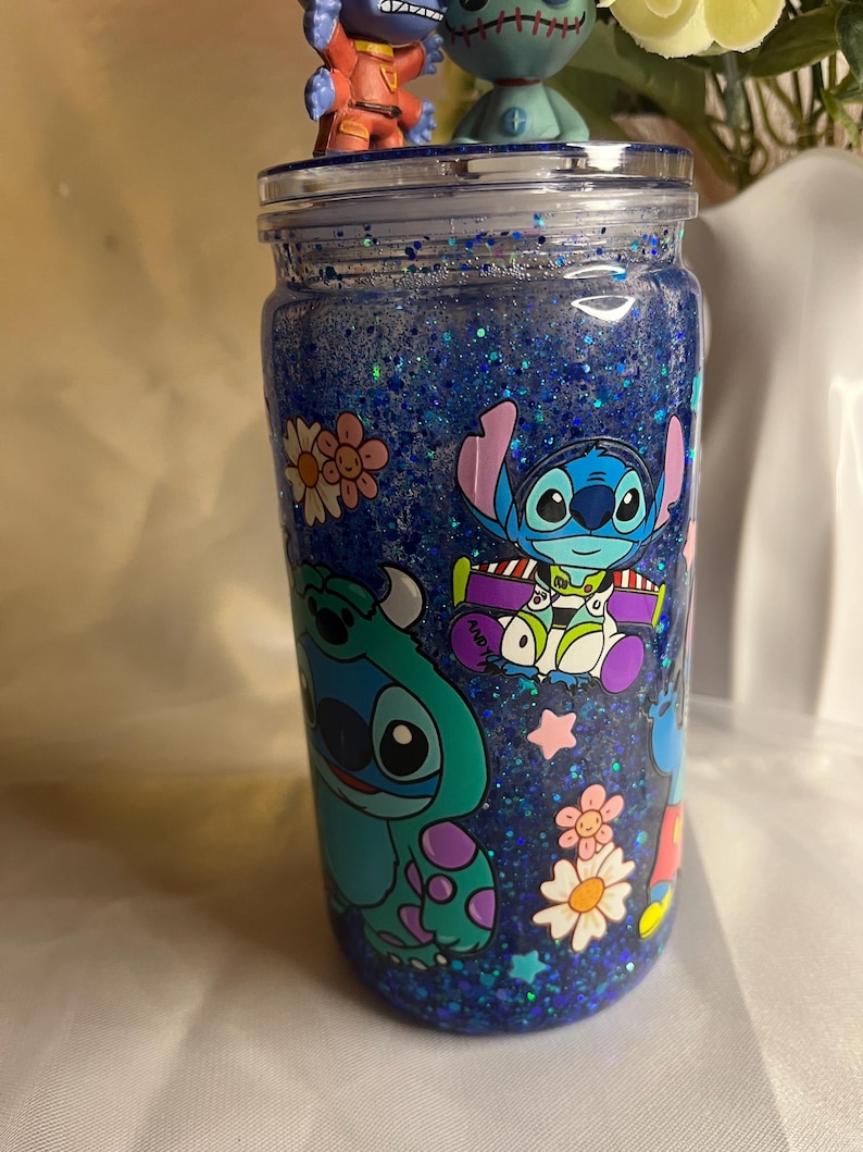 May include: A blue glitter tumbler featuring cartoon characters Stitch, Sulley, and Buzz Lightyear. The tumbler has a clear lid and is decorated with flowers and stars. Two character figurines sit on top.