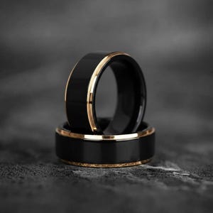 May include: Two rings with a polished black finish and a thin, metallic gold-toned inner band. One ring stands upright while the other rests horizontally beneath it, showcasing a smooth, reflective surface and a flat, wide band profile.