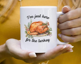 May include: White ceramic mug with the text "I'm just here for the turkey" and a watercolor illustration of a roasted turkey with herbs and flowers. The mug has a curved handle.