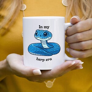 May include: White ceramic mug with a blue cartoon snake illustration and the text "In my herp era". The mug has a white handle. The mug is held by a person wearing a yellow top.