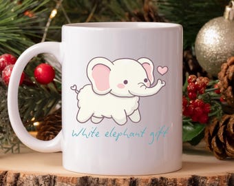 May include: White ceramic mug featuring a cute cartoon elephant with pink ears and a heart. The mug has the text "White elephant gift" in a light blue script. The mug is on a wooden surface with Christmas decorations in the background.