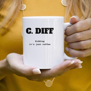 May include: White ceramic coffee mug with the text "C. DIFF" in bold black letters. Below the text, the mug reads "Kidding it's just coffee" in a smaller, black font. The mug is held by a person.