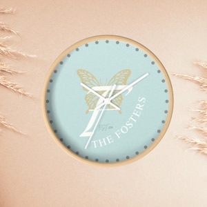 Può includere: Orologio da parete rotondo con quadrante azzurro con un disegno a farfalla dorata e la scritta "THE FOSTERS". L'orologio ha una cornice in legno e lancette bianche. Il quadrante include anche piccoli punti grigi sul bordo.