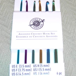 Może przedstawiać: Zestaw sześciu anodowanych haczyków do szydełkowania w różnych kolorach, w tym turkusowym, fioletowym, zielonym, złotym, niebieskim i ciemnozielonym. Haczyki są oznaczone rozmiarami amerykańskimi i odpowiadającymi im wymiarami w milimetrach: US E (3,5 mm), US F (3,75 mm), US G (4,5 mm), US H (5 mm), US I (5,5 mm) i US J (6 mm). Opakowanie zawiera również tekst "OOPS & THREADS" i "ANODIZED CROCHET HOOK SET".