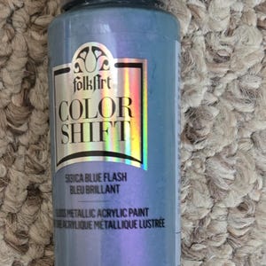 Może przedstawiać: Butelka farby akrylowej FolkArt Color Shift Blue Flash. Etykieta zawiera tekst "COLOR SHIFT" i "5131CA BLUE FLASH BLEU BRILLANT". Butelka zawiera 59 ml.