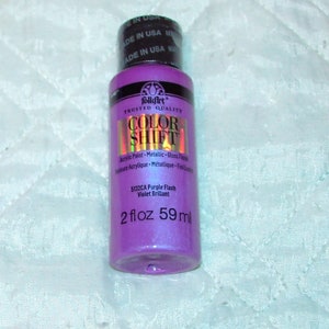 Może przedstawiać: Butelka farby akrylowej FolkArt Color Shift o pojemności 59 ml w odcieniu 5132CA Purple Flash Violet Brillant. Butelka została wyprodukowana w USA.