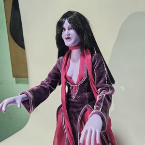 Puede incluir: Una muñeca con piel de color morado claro, cabello negro largo y liso, y labios rojos. La muñeca viste un vestido de terciopelo morado de manga larga con adornos plateados y una bufanda de tela rosa drapeada alrededor del cuello.