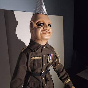 Puede incluir: Muñeca de estilo vintage con rostro bronceado, ojos negros y gorro de fiesta plateado. La muñeca viste uniforme militar verde oscuro con adornos dorados, cinturón negro y medalla. La muñeca tiene una ligera sonrisa.