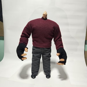 Puede incluir: Una figura de acción articulada con una cabeza pequeña y calva, que viste un suéter de manga larga acanalado color granate, pantalones gris oscuro y guantes negros sin dedos. La figura tiene brazos grandes y musculosos con dedos color carne.