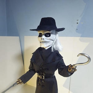 Puede incluir: Una muñeca de colección con rostro blanco y detalles oculares negros, que lleva un sombrero fedora negro y una gabardina larga negra de doble botonadura con cinturón. La muñeca tiene cabello blanco y un gancho de metal plateado.