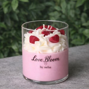 Puede incluir: Una vela rosa en un vaso transparente, cubierta con cera batida blanca y detalles rojos en forma de corazón. La vela tiene el texto "Love Bloom" y "by neha" impreso.