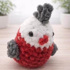 May include: A handmade, plush bird toy with a white head, red body, and gray accents. The bird has black button eyes and a heart-shaped red beak. The toy is made of soft, textured yarn.