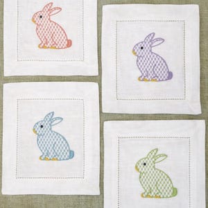 Pode incluir: Quatro bases de linho branco, cada uma bordada com um coelho de cor diferente: rosa, roxo, azul e verde. Os coelhos têm detalhes amarelos e estão centrados nas bases quadradas. Cada base tem uma borda pontilhada.