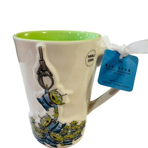 Pu&ograve; includere: Una tazza in ceramica color crema con interno verde brillante, caratterizzata da un design in rilievo di alieni verdi a tre occhi di Toy Story sollevati da un artiglio. Un'etichetta blu attaccata con un nastro bianco riporta la scritta RAE DUNN ARTISAN COLLECTION BY MAGENTA.