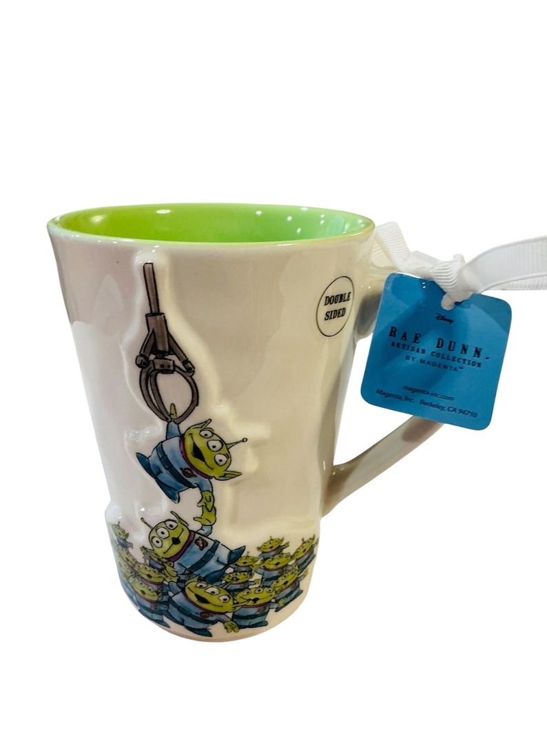 Pu&ograve; includere: Una tazza in ceramica color crema con interno verde brillante, caratterizzata da un design in rilievo di alieni verdi a tre occhi di Toy Story sollevati da un artiglio. Un'etichetta blu attaccata con un nastro bianco riporta la scritta RAE DUNN ARTISAN COLLECTION BY MAGENTA.