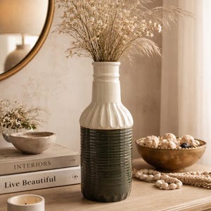 May include: A decorative vase with a white top and a dark green base, filled with dried flowers. The vase is on a wooden table, alongside a bowl, a candle, and books. A gold-framed mirror is in the background.