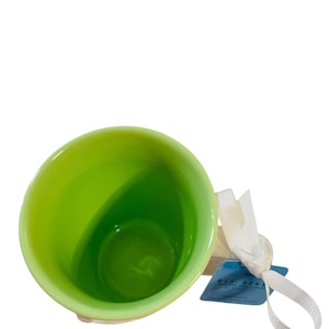 Pu&ograve; includere: Una tazza in ceramica con interno verde lime brillante ed esterno bianco. Un nastro bianco &egrave; legato attorno al manico e una piccola etichetta rettangolare blu con il testo Rae Dunn &egrave; attaccata al nastro vicino al manico della tazza.