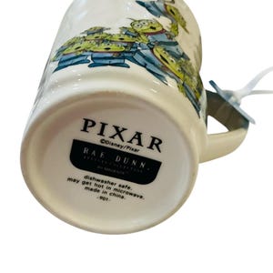 Pu&ograve; includere: Una tazza in ceramica bianca con una grafica raffigurante diversi alieni verdi a tre occhi. La base riporta il testo PIXAR, Disney/Pixar, RAE DUNN, ARTISAN COLLECTION BY MAGENTA, dishwasher safe, may get hot in microwave, made in china, -901-.