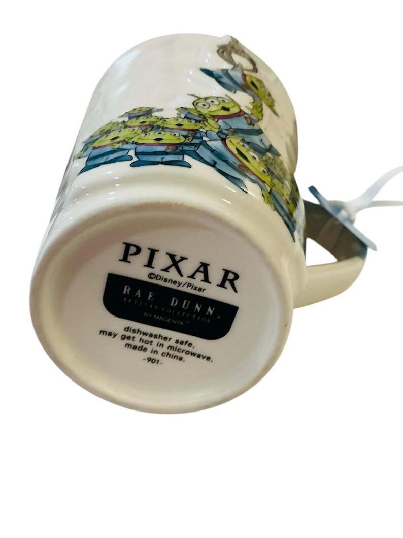 Pu&ograve; includere: Una tazza in ceramica bianca con una grafica raffigurante diversi alieni verdi a tre occhi. La base riporta il testo PIXAR, Disney/Pixar, RAE DUNN, ARTISAN COLLECTION BY MAGENTA, dishwasher safe, may get hot in microwave, made in china, -901-.