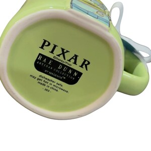 K&ouml;nnte beinhalten: Eine gr&uuml;ne Keramiktasse mit dem Pixar-Logo und dem Text Rae Dunn Artisan Collection. Die Tasse hat einen wei&szlig;en Henkel und ein Design am Rand. Der Text "sp&uuml;lmaschinenfest" und "made in china" sind ebenfalls sichtbar.