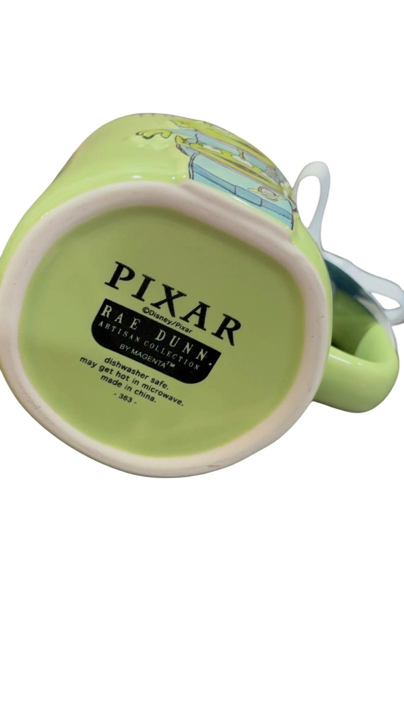 K&ouml;nnte beinhalten: Eine gr&uuml;ne Keramiktasse mit dem Pixar-Logo und dem Text Rae Dunn Artisan Collection. Die Tasse hat einen wei&szlig;en Henkel und ein Design am Rand. Der Text "sp&uuml;lmaschinenfest" und "made in china" sind ebenfalls sichtbar.