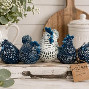 May include: A set of five ceramic chicken figurines arranged in a row on a white wooden tray. Four chickens are dark blue with white textured details, while the central chicken is white with blue accents. A brown paper tag labeled KITCHEN is attached.