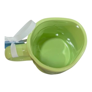K&ouml;nnte beinhalten: Eine hellgr&uuml;ne Keramiktasse mit abgerundeter Form und Henkel. Die Tasse hat eine gl&auml;nzende Oberfl&auml;che und ein wei&szlig;es Band am Henkel. Das Innere der Tasse ist in einem helleren Gr&uuml;nton gehalten.