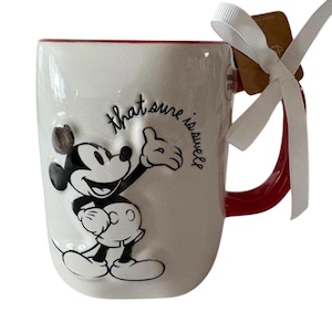 May include: White ceramic mug featuring Mickey Mouse in black and white, with a red handle and rim. The mug has the phrase "that sure is swell" in black script. A white ribbon and tag are attached to the handle.