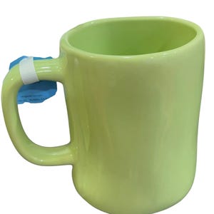 K&ouml;nnte beinhalten: Eine hellgr&uuml;ne Keramiktasse mit einem abgerundeten Henkel. Die Tasse hat eine glatte, gl&auml;nzende Oberfl&auml;che und ein schlichtes, modernes Design. Die Tasse ist leer und bereit, mit einem Getr&auml;nk gef&uuml;llt zu werden. Ein wei&szlig;es Etikett ist am Griff befestigt.