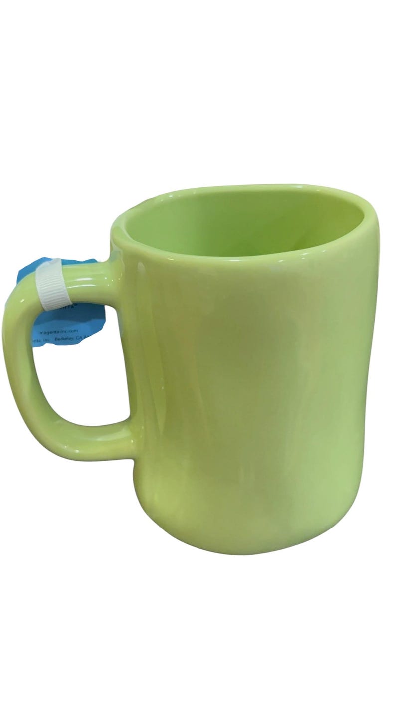 K&ouml;nnte beinhalten: Eine hellgr&uuml;ne Keramiktasse mit einem abgerundeten Henkel. Die Tasse hat eine glatte, gl&auml;nzende Oberfl&auml;che und ein schlichtes, modernes Design. Die Tasse ist leer und bereit, mit einem Getr&auml;nk gef&uuml;llt zu werden. Ein wei&szlig;es Etikett ist am Griff befestigt.
