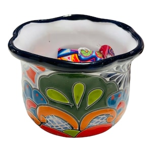 May include: A hand-painted ceramic pot with a scalloped rim and a dark blue border. The pot features a vibrant floral design in green, orange, blue, and red. The interior is white, and the pot contains a small, colorful object.