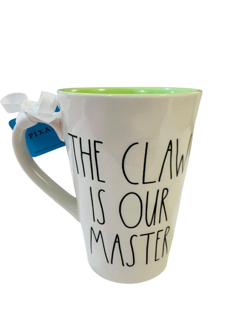 Pu&ograve; includere: Una tazza in ceramica bianca con interno verde chiaro. L'esterno presenta un testo nero scritto a mano che recita THE CLAW IS OUR MASTER. Un nastro bianco &egrave; legato al manico e un'etichetta blu con la parola PIXAR &egrave; attaccata al manico.