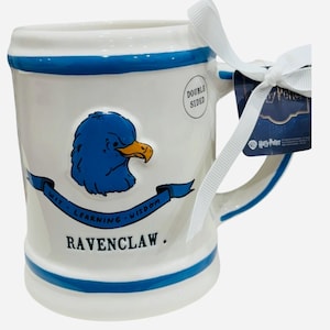May include: White ceramic mug with blue accents and a Ravenclaw crest. The mug features a blue eagle with a yellow beak, the words "Wit, Learning, Wisdom" and "Ravenclaw." A white ribbon and a Harry Potter tag are attached.