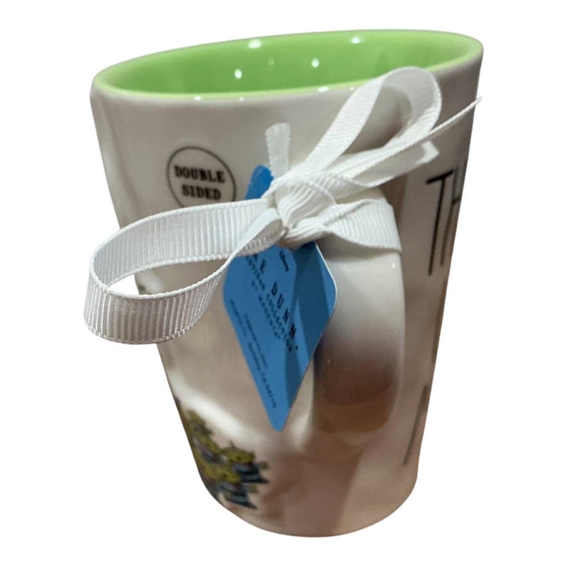 Pu&ograve; includere: Una tazza in ceramica bianca con interno verde lime brillante. La tazza presenta un nastro bianco legato attorno al manico, che sostiene un'etichetta blu con la scritta Rae Dunn. Un piccolo adesivo circolare sul lato riporta la scritta Double Sided.
