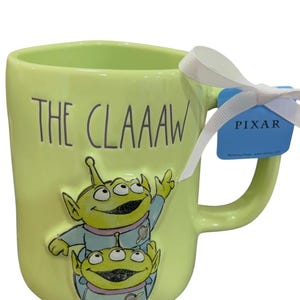 K&ouml;nnte beinhalten: Eine hellgr&uuml;ne Keramiktasse mit dem Text "THE CLAAAW" und einer Illustration von zwei gr&uuml;nen Aliens. Die Tasse hat ein wei&szlig;es Band und ein blaues Pixar-Etikett am Griff. Ein Sammlerst&uuml;ck.