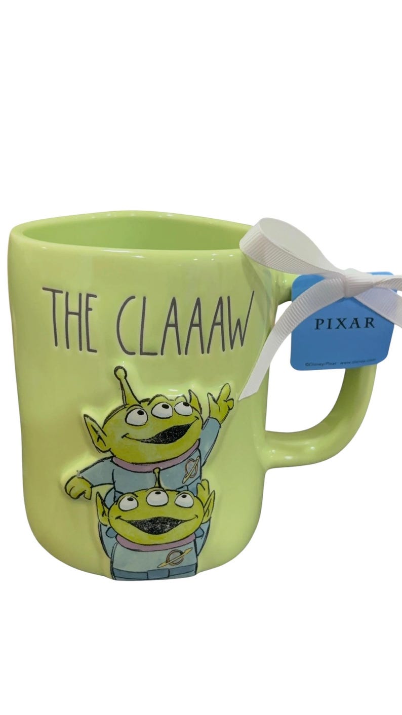K&ouml;nnte beinhalten: Eine hellgr&uuml;ne Keramiktasse mit dem Text "THE CLAAAW" und einer Illustration von zwei gr&uuml;nen Aliens. Die Tasse hat ein wei&szlig;es Band und ein blaues Pixar-Etikett am Griff. Ein Sammlerst&uuml;ck.