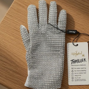 May include: A silver glove covered in small, sparkling embellishments. The glove has a white cuff and is paired with a tag that reads "Thriller" and includes maintenance instructions. The glove is resting on a wooden surface.