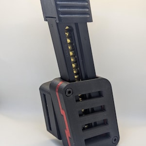 Puede incluir: Soporte de cargador negro con un acento rojo. El cargador es negro con balas doradas visibles. El texto "TAURUS TX" está impreso en el lateral del cargador.