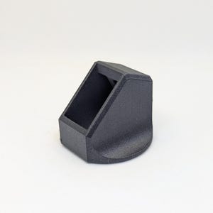 Puede incluir: Un soporte angular negro, impreso en 3D, con una abertura rectangular y una base curva. El soporte está diseñado para sostener un objeto en ángulo. El objeto está hecho de un material negro mate.