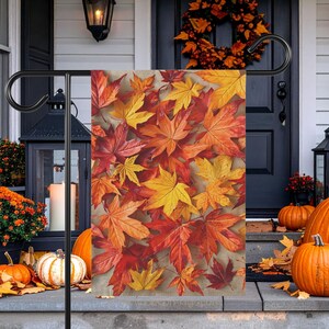 以下が含まれることがあります： 黄色、オレンジ、深紅の色合いの秋のカエデの葉が重なり合う鮮やかな模様が特徴の長方形のガーデンフラッグ。黒い金属製のスタンドに掲げられており、密集した色とりどりの秋の紅葉のデザインが描かれています。